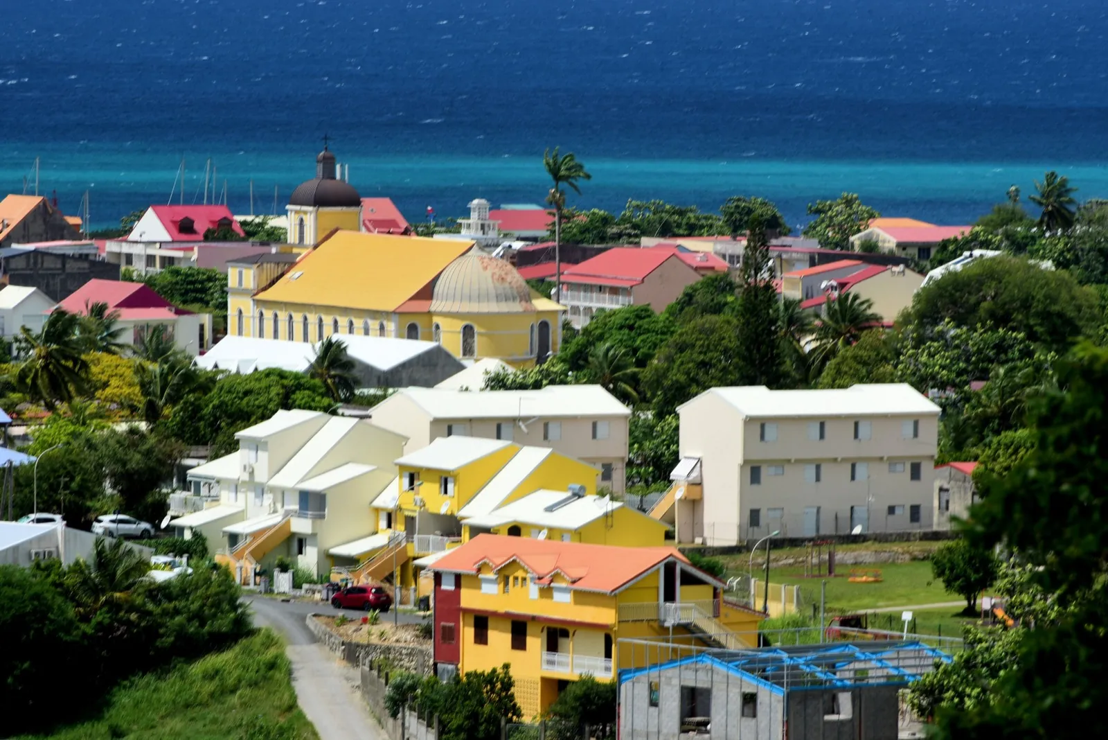 Grand-Bourg vu des hauteurs : maisons colorées, église au toit jaune et à la coupole grise, et la mer turquoise à l'horizon